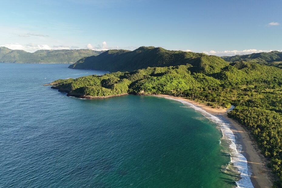 Aerial view of Playa Cosón, with blue water, a curved sandy shore and green hills.